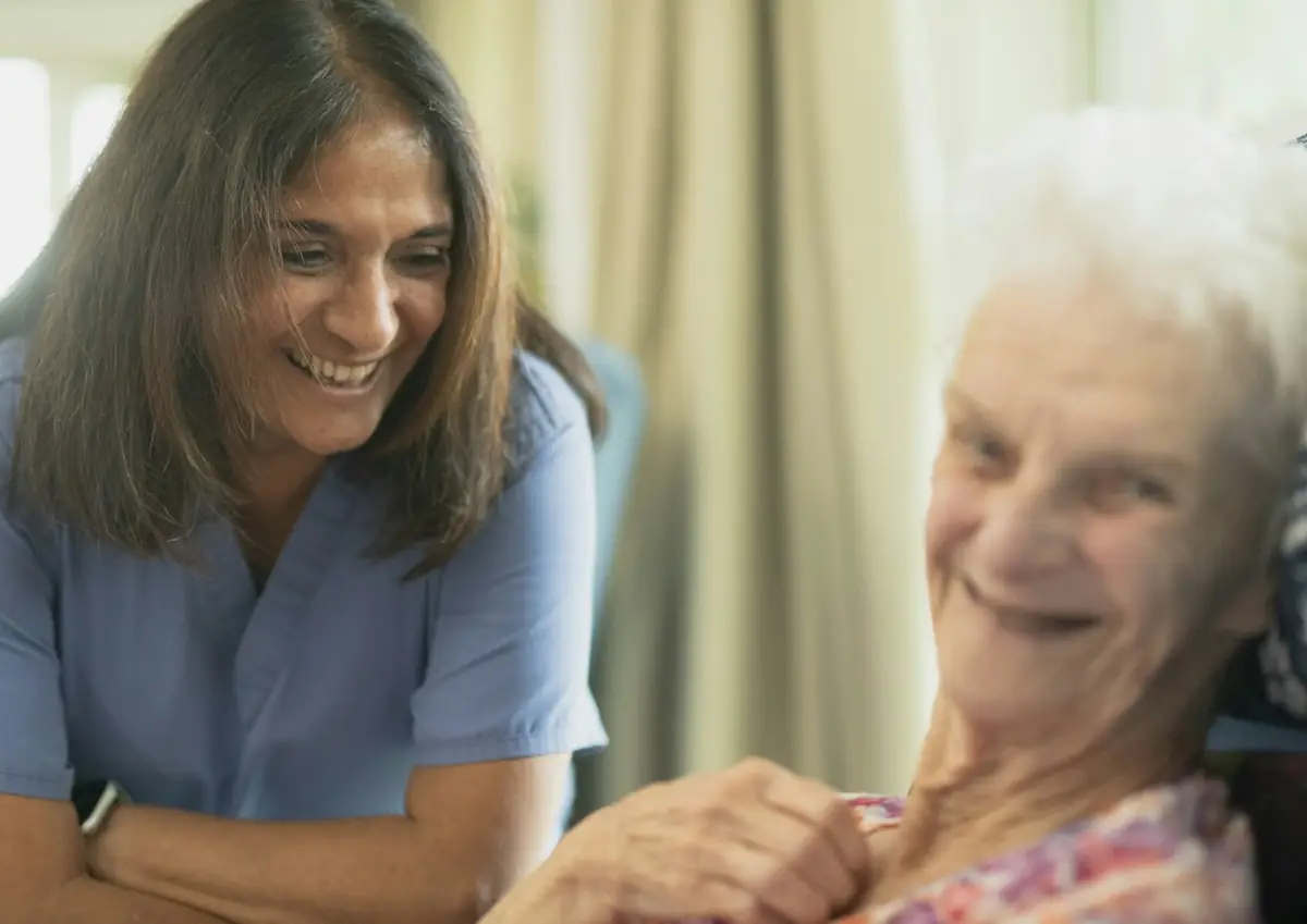 A smiling nurse in blue scrubs talking with an older woman at home